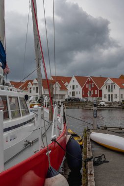 Bulutlu bir gökyüzünün altında kırmızı ve beyaz bir yelkenli dinleniyor. Turuncu kiremitli beyaz ve kırmızı ahşap evler fiyort köyünün rıhtımında..