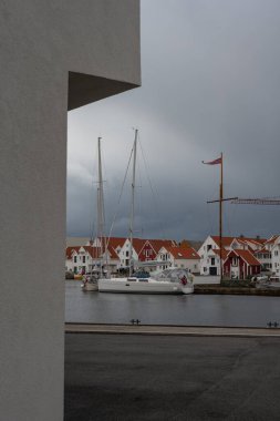 Beyaz bir yelkenli Norveç limanında turuncu kiremitli çatıları olan kırmızı ve beyaz ahşap evlerin yanına yanaştı. Gökyüzü bulutlu ve ön planda asfaltlı rıhtım.