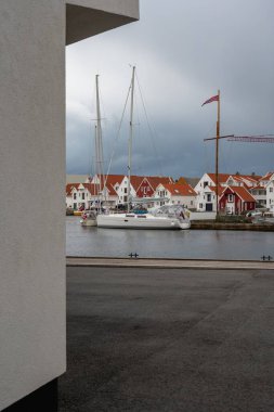Beyaz bir yelkenli, kırmızı ve beyaz ahşap evlerin yakınındaki sakin bir limanda dinleniyor. Bir Norveç bayrağı, bir vinç ve bir beyaz bina rıhtım sahnesini çerçeveliyor..