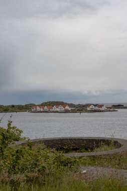 Norveç 'te, sakin sularla çevrili, beyaz evleri ve kırmızı çatıları olan kayalık bir ada. Ön planda dairesel beton yapıya sahip çimenli bir alan var..