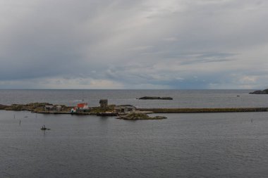 Sakin sularda küçük bir Norveç yerleşimi, kırmızı çatılı bir bina, taş bir rıhtım, kayalık adalar, ve açık denizli bulutlu bir gökyüzü..