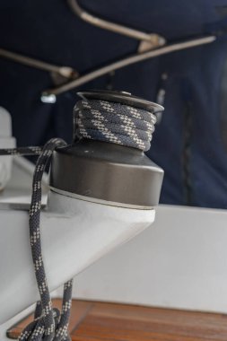 Beyaz bir yüzeye sarılı halatla bir yelkenli vincin detaylı bir görüntüsü, koyu mavi bir gölgelik ve kısmen görünür ahşap bir güverteye kurulmuş..