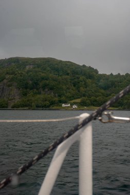 Bir yelkenli Norveç 'te bir fiyort çerçeveliyor, yeşil bir yamaç, kıyıya yakın iki beyaz ev, ve arka planda bulutlu bir gökyüzü..