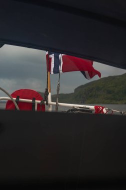 Norveç bayrağı, korkuluğu ve kırmızı can yeleği olan bir tekne. Bulutlu bir gökyüzünün altında yemyeşil bir yamaç yükseliyor..