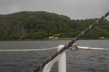 Norveç 'te halatlarla ve fiyortlarla donatılmış bir yelkenli ön planda. Sahile yakın iki beyaz evi olan yeşil bir yamaç arka planda yükseliyor..