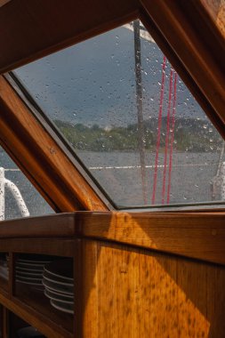 İçi yağmurla kaplı pencere, kırmızı ipler, istiflenmiş tabaklar ve Norveç 'te sakin bir fiyort ve bulutlu bir gökyüzü manzaralı ahşap bir yelkenli kabini..