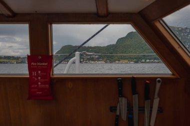 Norveç fiyordunu gösteren bir teknenin içinden görüntü, yeşil tepelerde renkli evler ve yağmurlu bir pencere. İç ayrıntılar arasında itfaiye battaniyesi ve bıçaklar var..