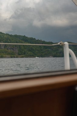 Ön planda bir teknenin korkuluğu ve ahşap bir yapı var. Arka planda kayalık çıkıntıları, sakin suyu ve küçük uzak tekneleri olan yeşil bir yamaç yer alıyor..