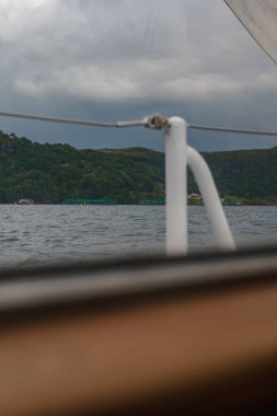 Ön planda, yemyeşil bir kıyı şeridi, dağınık binalar, deniz ürünleri kalemleri ve bulutlu gökyüzü altında sakin bir su bulunan bir yelkenli yelkenlisi görünüyor..