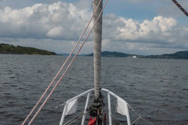 Yelkenlinin pruvası görünür bir direk ve donanımlı Norveç 'te sakin bir fiyortta ilerliyor, yeşil tepeler, dağlar ve uzak bir yerleşim yeriyle çevrili..