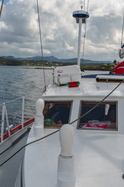 Beyaz güvertesi ve kabini olan küçük bir tekne engebeli bir kıyı şeridi yakınlarında sakin sularda yol alır. Sağda bulutlu bir gökyüzünün altında bir deniz feneri duruyor..