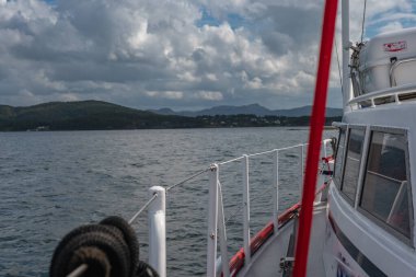 Norveç 'te sakin bir fiyortta seyreden bir tekne, bir cankurtaran botu, orman tepeleri, dağınık evler ve parçalı bulutlu bir gökyüzü..