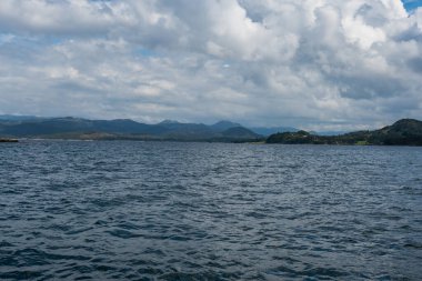 Norveç 'te sakin bir fiyort, sakin sular, ormanlık kıyılar, açık yeşil alanlar, ve parçalı bulutlu bir gökyüzünün altında tepeler ve dağlardan oluşan bir zemin..