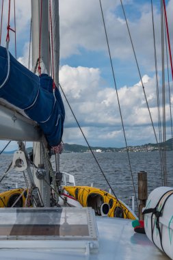 Mavi yelkenli ve sakin sularda sarı sallı bir yelkenli güvertesi. Kıyı şeridi bulutlu bir gökyüzünün altında dağınık binalar ve yeşil tepelere sahiptir..