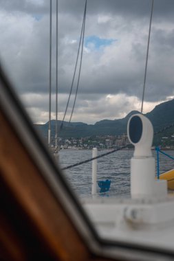 Ön planda yelkenli güvertesi ve teçhizat var. Arka planda yeşil tepelerin altında bir kıyı kasabası var. Parçalı bulutlu bir gökyüzünün altında..