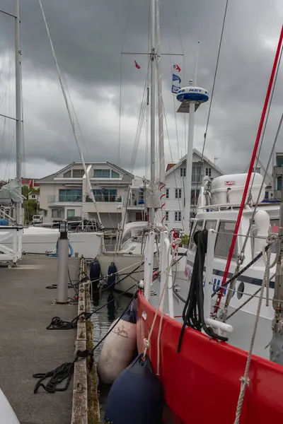 Yelkenliler Norveç 'te beton bir rıhtıma demirlediler ve ön planda kırmızı bir tekne vardı. Geniş pencereli beyaz binalar, bulutlu sahil şeridi..