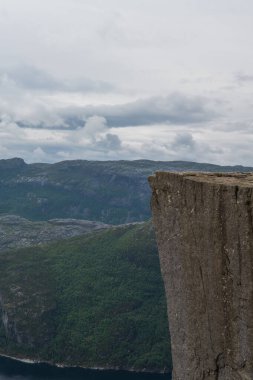 İkonik Preikestolen uçurum Norveç 'te Lysefjord' un üzerinde yükselir, dik kaya yüzeyleri, yemyeşil tepeler, engebeli dağlar ve bulutlu bir gökyüzü.