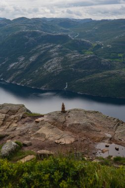 Norveç 'te Lysefjord' un kayalık bir çıkıntıda bir cairn, uzak bir şelale, engebeli dağlar ve bulutlu bir gökyüzünün altında sakin yansıtıcı sulara sahip görüntüsü.