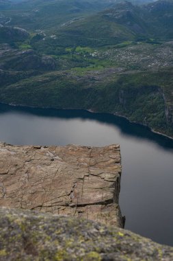 Preikestolen 'in engebeli düz tepesinin manzarası, Lysefjord' un sakin suları ve arka planda yeşil tepeler, küçük yerleşim yerleri ve doğal manzarayla.