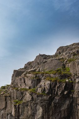 Yeşil bitki örtüsü ve tepesinde bir kişi olan yüksek gri bir uçurum Norveç 'in Preikestolen veya Lysefjord bölgesinde açık mavi bir gökyüzüne karşı uzanıyor..