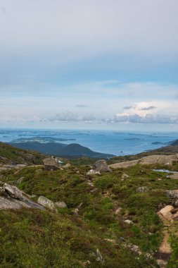 Yeşil bitki örtüsü ile kayalık arazi, dolambaçlı bir yol, ve küçük adaları olan fiyort, tepelerle çevrili ve Norveç 'te parçalı bulutlu bir gökyüzü.