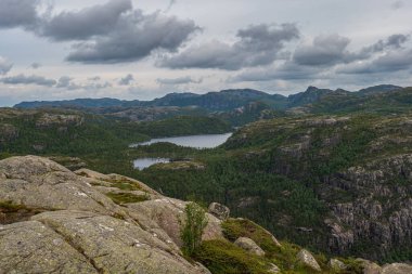 Ön planda engebeli kaya oluşumları, yoğun yeşil ormanlar, dingin bir göl ve Lysebotn, Norveç yakınlarındaki bulutlu bir gökyüzünün altında dalgalanan tepeler..