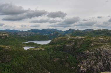 Norveç 'in Lysebotn yakınlarındaki engebeli arazi, yemyeşil ormanlar, dingin bir göl, kayalık uçurumlar ve dağınık bulutlarla kaplı bulutlu bir gökyüzü..