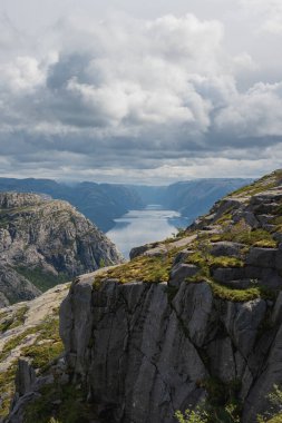 Norveç 'teki Lysefjord manzarası, sarp kayalıklar ve sarp kayalıklarla çevrili. Yeşil bitki örtüsü yukarıdaki dramatik bulutlu gökyüzü ile çelişiyor.