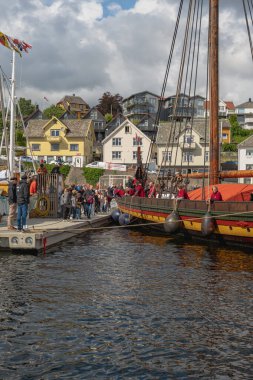 Tahta gövdeli ve uzun direkli bir Viking gemisi marinaya yanaşmış, etrafı insanlarla çevrili. Yakınlarda renkli evleri olan bir yamaç köyü görülüyor..