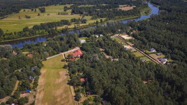 Polonya 'nın Nowy Lubiel yakınlarındaki kırsal bir bölgenin hava görüntüsü, kıvrımlı bir nehir, orman arazisi, açık alanlar ve kırmızı çatılı binalar kümesini gösteriyor..