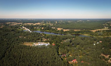 Rüzgarlı bir nehri olan panoramik bir hava manzarası, açık mavi gökyüzünün altında kırmızı çatılı evler, açık araziler ve tarım arazileri olan küçük bir yerleşim yeri..
