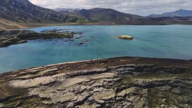 Işıl ışıl mavi bir göl, çimenli bir ada, karanlık kıyı şeritleri ve yeşil bitki örtüsü, İzlanda 'nın doğal arazisini gösteriyor..