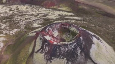 Drone volkanik krateri kırmızımsı renklerle kaplıyor, yosun kaplı lav tarlaları, toprak yollar, mavi bir göl ve İzlanda 'da konik bir dağ..