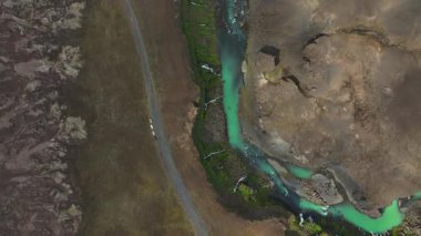 Turkuaz bir nehir, yosun kaplı kayalar ve park halindeki araçlarla çevrili İzlanda engebeli arazisinden akar..