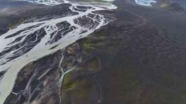 İnsansız hava aracı volkanik ovalarda örülmüş nehirleri süpürüyor. Arka planda karlı dağlar, göller ve yeşil bitkiler var..