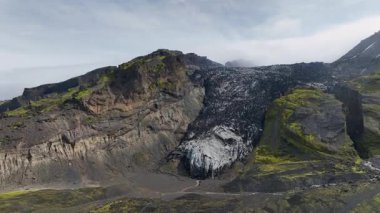 İnsansız hava aracı İzlanda 'daki bir buzulun üzerinden yükseliyor, buzlu yüzeyler, yosun kaplı yamaçlar ve dramatik bir dağ manzarasındaki dik kayalıklar ortaya çıkıyor..