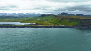 Reynisfjara 'nın kara kumsal manzarası, yemyeşil uçurumlar, kırsal yollar ve bulutlu bir gökyüzünün altındaki karlı dağlar..