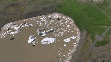 İzlanda 'da yeşil çimenli araziyle çevrili çamurlu kahverengi sularda sürüklenen beyaz buzdağları olan bir buzul gölünün hava görüntüleri..