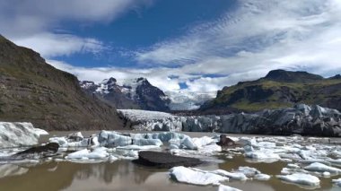 İnsansız hava aracı, engebeli dağlar ve parçalı bulutlu bir gökyüzünün altındaki yeşil yamaçlarla çevrili yüzen buzdağlarıyla gölün içine akan bir buzulu yakalar..