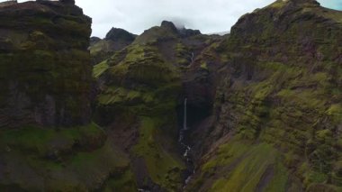 İnsansız hava aracı İzlanda 'da yosun kaplı bir kanyonun üzerinden yükselir. Dar bir şelale, sivri kayalıklar ve kısmen bulutlarla gizlenmiş bir buzulu ortaya çıkarır..