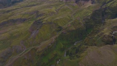 İnsansız hava aracı, dik uçurumlar, yosun kaplı yamaçlar, çağlayan şelaleler ve aşağıdaki dolambaçlı bir nehir ile verimli bir İzlanda kanyonunda süzülüyor..