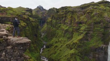İnsansız hava aracı İzlanda 'da yemyeşil bir kanyonda süzülüyor, yosun kaplı uçurumlar, dolambaçlı bir nehir ve bulutlu bir gökyüzünün altındaki uzun bir şelale ortaya çıkıyor..