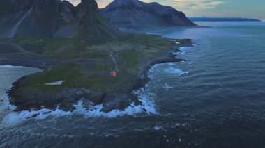İzlanda 'da yumuşak akşam gökyüzünün altında dalgalı dağlar ve okyanus dalgaları ile kayalık bir çıkıntıda turuncu bir deniz fenerinin drone görüntüsünü süpürüyor..