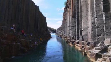 Turkuaz bir nehir bazalt sütunlarıyla dar bir kanyondan akar ve küçük bir şelaleye gider. Video nehri akıntıya karşı parlak bir gökyüzünün altında takip ediyor..