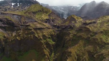 İnsansız hava aracı İzlanda 'da bir kanyonun üzerinden geçiyor, yosun kaplı uçurumlar, karanlık bir çakıl yatağı ve engebeli araziye yakın küçük beyaz bir araç ortaya çıkarıyor..