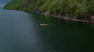 Lysebotn 'da sakin bir fiyort manzarası, dik orman uçurumları, sakin yeşil su, ve bulutlu bir gökyüzü altında kürek çeken yalnız bir kano..