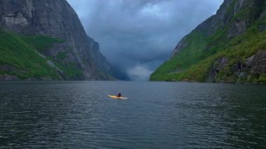 Norveç 'teki Lysebotn Fjord' un sakin bir hava manzarası. Yeşil yamalı yüksek uçurumlar durgun suyun üzerinde yükseliyor, bir şelale ve bulutlu gökyüzü.