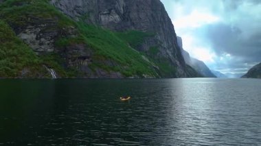 İnsansız hava aracı Lysebotn 'daki fiyortta yeşil yamalı sarp kayalıkları, sakin yansıtıcı suyu ve bulutlu gökyüzünü aydınlatan güneş ışığını gösterir..
