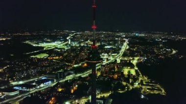 Litvanya 'daki Vilnius TV Kulesi' nin geceleri kırmızı ışıklarla parlayan görüntüsü. Çevresindeki şehir manzarasında canlı caddeler ve binalar yer alıyor..