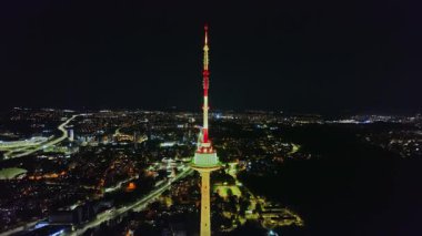 Gece Vilnius TV Kulesi 'nin havadan görünüşü, kırmızı ve yeşille aydınlatılmış. Kamera düzgün hareket ediyor, kuleyi ve yanan şehri gösteriyor..